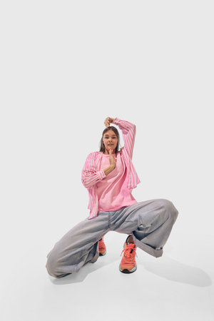 Woman kneels playfully, while dancing energetically in pink and loose denim, against white studio...