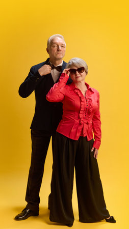 Sophisticated pair, man in tailored suit standing beside woman in modern attire and shades, posing confidently against yellow studio background. Concept of mature people in modern lifestyle, fashion.の写真素材