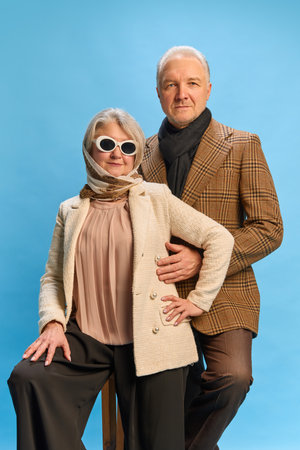 Duo sitting side by side, woman in stylish attire holding white sunglasses, man in retro outfit with relaxed expression, against blue studio background. Concept of mature people and fashion. Adの写真素材