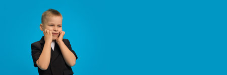 Young boy in black blazer, white shirt, cupping face with hands, smiling shyly while standing at side against bright blue background. Concept of childhood, beauty, fashion, style.の写真素材