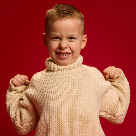 Portrait of smiling boy in beige sweater, holding arms bent, flexing muscles with confident grin...