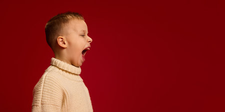 Boy widely open mouth, turning sideways, as he shouting loudly with aggression, dressed in knitted sweater against deep red background. Concept of childhood, fashion, style.の写真素材
