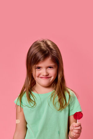 Portrait of playful child holding red lollipop, looking mischievous with small grin against pink studio background. Concept of healthcare, protection, immunity, prevention.の写真素材