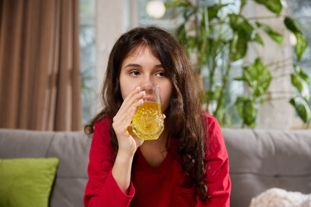 Woman in red pajama holds small white packet close to camera while sitting on couch with green...