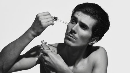 Man with smooth skin, applying serum with dropper against white background in black-and-white...