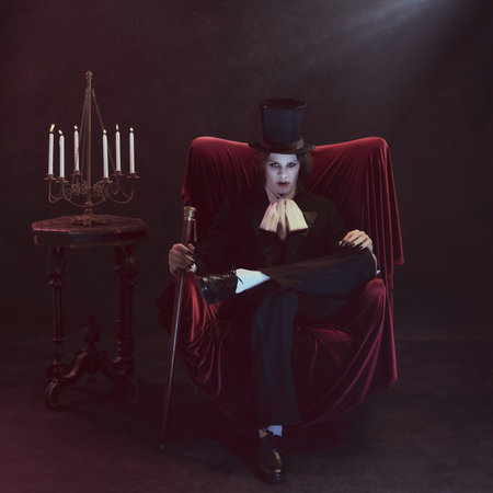 Terrifying man, vampire with sharp fangs, dressed in strict vintage suit and cylinder, sits in...