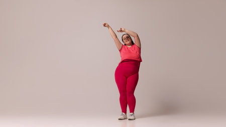 Fat woman in pink top, red leggings, and white sneakers stretching arms upward with relaxed expression, against beige studio background. Concept of beauty, fitness, movement, dance and music.の写真素材