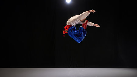 Male dancer executing backward jump, dressed in traditional embroidered shirt, blue trousers, and...