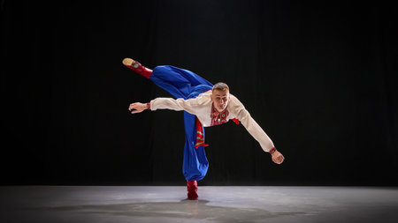 Dancer in white embroidered shirt and blue trousers balances gracefully on one foot, leaning forward...
