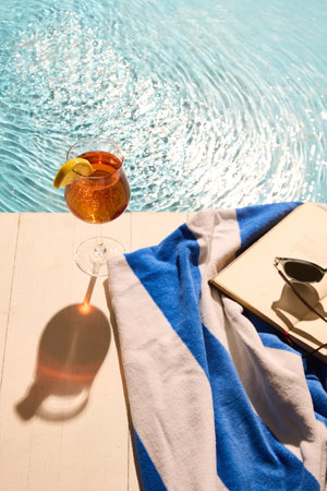 Summer drink with lemon wedge, striped towel, book, and black sunglasses on deck by sunlit pool, evoking peaceful relaxation and warm holiday vibes. Concept of summertime recreation, tourism. Adの写真素材