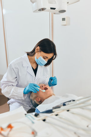 Female doctor leaning over elderly woman in dental chair, performing oral examination with tools at bright modern dental office. Concept of healthcare and pensioners, medicine.の写真素材