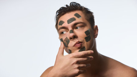 Photo of man with wet, brunette hair, doing facial procedures, applying black clay mask on skin against white studio background. Concept of selfcare and love, natural beauty, cosmetology.の写真素材