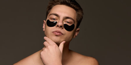 Calm and confident young handsome man with black hydrogel patches posing, holding hand on chin...