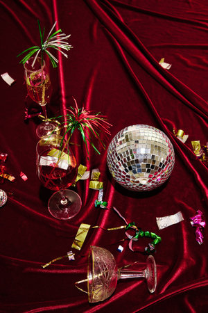Disco ball, cocktail glasses with golden confetti, and spilled drinks on red velvet fabric, evoking...