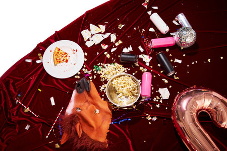 Spilled popcorn, broken plates, empty pizza box, and horse mask on red velvet, visualizing surreal aftermath of crazy night with alcohol and snacks. Concept of alcohol, hangover, afterparty mood.の写真素材