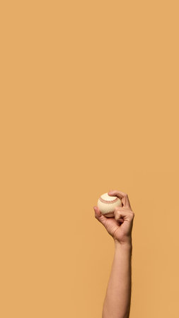 Hand holding baseball high, fingers gripping seams with precision, symbolizing control and readiness for action against warm-toned background. Concept of sport equipment, control, strategy.の写真素材