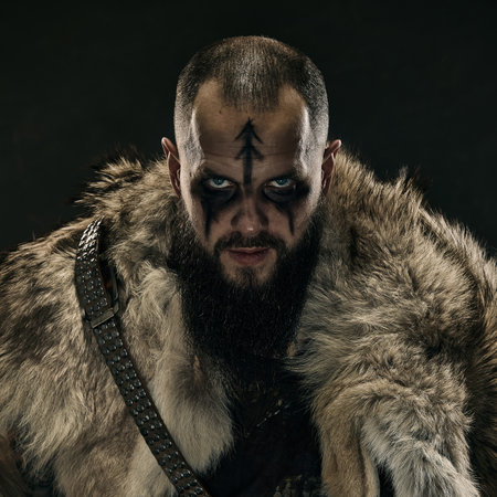 Close-up portrait of Viking warrior with war paint, piercing blue eyes, and fur cloak, staring menacingly against dark background. Concept of medieval portrait, Norse warrior spirit, traditions.の写真素材