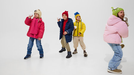 Group of children, wearing colorful winter jackets and hats, standing ready with snowballs, smiling and enjoying playful winter fun against white studio background. Concept of winter vacation.の写真素材