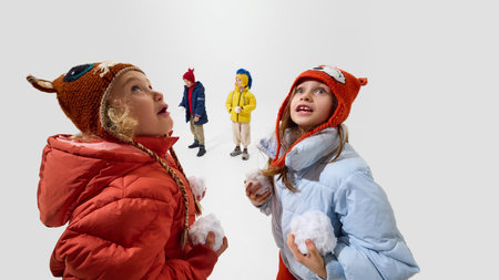 Kids, dressed warm winter jackets and knitted hats, holding snowballs, looking excited and playful, preparing for winter game against white background. Concept of childhood, winter vacation, carefree.の写真素材