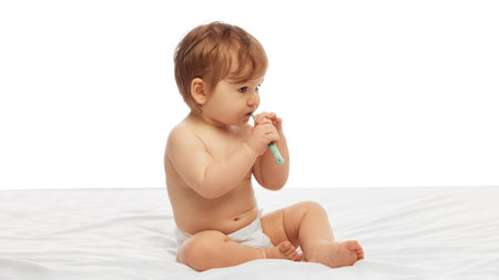 Baby in diaper sitting on soft white bed, and chewing toothbrush while holding it with both hands against white background. Concept of childhood, hygiene, health, early habits.の写真素材