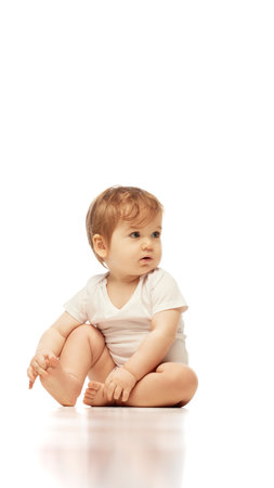 Baby, dressed white onesie, sitting cross-legged while looking sideways with focused expression against white background. Concept of childhood, born and growth, awareness, observation, early cognitionの写真素材