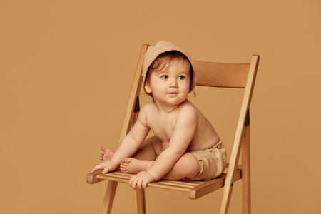 Happy little baby sitting on wooden chair, wearing soft cap and shorts, with playful expression...
