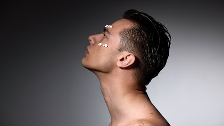 Man tilting head with white pills aligned on forehead and cheek, gazing into distance against dark...