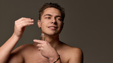 Man applying serum with dropper to cheek while holding glass bottle, smiling softly, looking to distance against brown studio background. Concept of selfcare and love, natural beauty, cosmetology.の写真素材