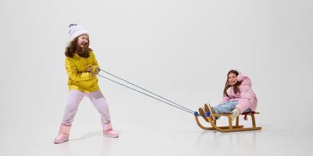 Girl pulling sled with friend, smiling, dressed in yellow winter coat, pink boots, having fun against white studio background. Concept of winter vacation, friendship, .の写真素材