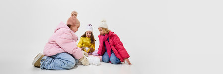 Banner. Three girls in colorful winter outfits sitting on floor, rolling snowballs and having fun together against white studio background. Concept of winter vacation, friendship, adventure.の写真素材