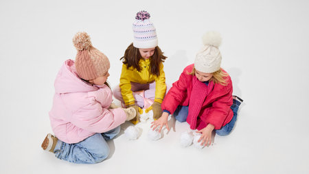 Three girls in cozy winter coats sitting together, laughing and building snowballs while enjoying winter fun against white studio background. Concept of winter vacation, friendship.の写真素材