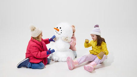 Little girls putting details on snowman, adjusting arms and facial features, enjoying winter play against white studio background. Concept of emotions, vacation, winter time, game.の写真素材