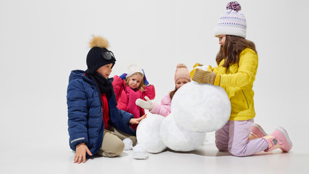 Boy and three girls dressed warm winter clothes reacting to snowman falling, girl in yellow jacket trying to stabilize, others expressing surprise. Concept of emotions, vacation, winter time, game.の写真素材