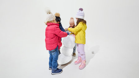 Group of children dressed warm winter clothes standing together building snowman, teamwork creating joyful winter scene against white background. Concept of vacation, winter time, game.の写真素材