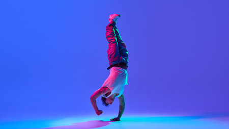 Dynamic image of flexible young guy in jeans and tank top dancing break dance against gradient blue purple background in neon light. Concept of dance, street style, creativity, youth culture, energyの写真素材
