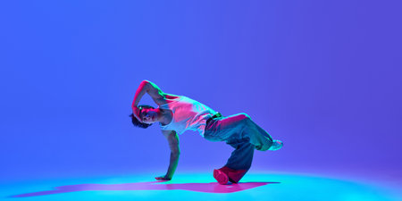 Skilled young dancer performing breathtaking breakdance tricks with against gradient blue purple background in neon light. Concept of hip-hop culture, modern street style, creativity, and movement.の写真素材