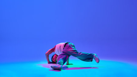 Athletic young man in motion, mid-air breakdance spin against neon blue purple background,...