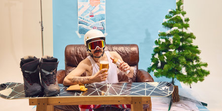 Man in ski goggles enjoying beer and hot dog, sitting on leather couch at home with Christmas tree in background. Concept of relaxation, enjoyment, leisure, winter vacationの写真素材