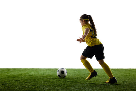 Determined child athlete in soccer gear running with ball on green field, isolated on white background with grassy surface. Concept of sport, childhood, motion, competition, training, activity.の写真素材