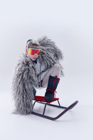 Young woman posing on sled in oversized shaggy fur coat, ski goggles, and moon boots against white...