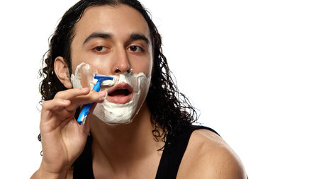 Young man with curly wet hair doing morning routine, shaving face with blue razor, thick foam covering skin against white studio background. Concept of male grooming, shaving, skincare, self-careの写真素材