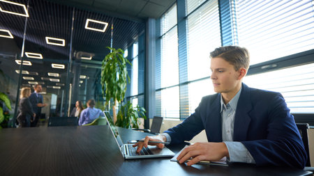 Focused young businessman typing on laptop at dark wooden table in sleek, glass-walled office, with...