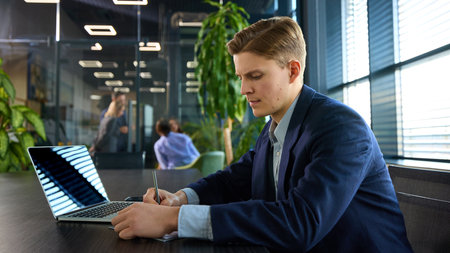 Attentive young businessman writing in notebook at desk with laptop open, maintaining structured approach in professional setting. Concept of planning, organization, efficiency, career development.の写真素材