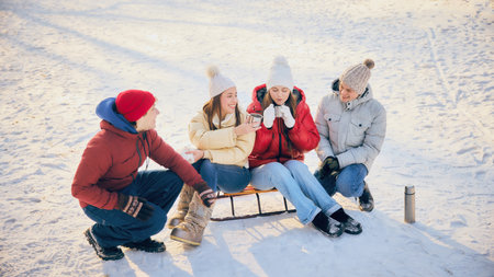 Group of friends, men and women, dressed comfy winter clothes, sitting in snow, sharing hot drinks,...