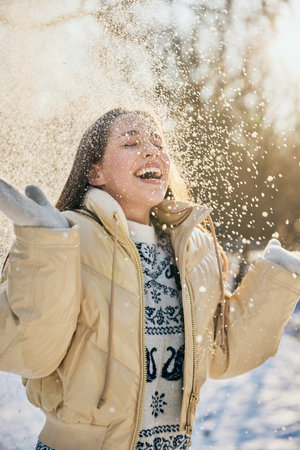 Excited young woman in beige jacket and cozy winter gloves throws snow in air, laughing in sunny winter park. Concept of winter, vacation, human emotions, nature, leisure time.の写真素材