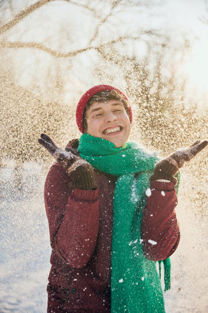 Young man in red hat and green scarf with snow-covered jacket smiles warmly in winter scenery. Concept of winter, vacation, human emotions, nature, leisure time.の写真素材