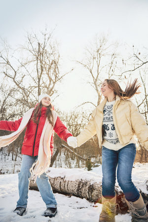 Two young women, dressed cozy winter outfits, holding hands, smiling as they play in snow-covered landscape. Concept of winter, vacation, human emotions, nature, leisure time.の写真素材