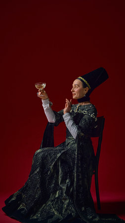 Elder noblewoman in renaissance attire holding a glass, smiling gently while raising it against a deep red background. Concept of art, noble times, joy, elegance, vintage celebration.の写真素材
