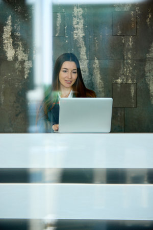Businesswoman focused on laptop screen while sitting at reception desk, glass reflections blending with office interior. Concept of technology, efficiency, workplace innovation.の写真素材
