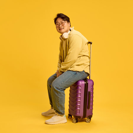 Cheerful Asian man, dressed casually, sitting on huge travel bag, ready to new adventures against sunny yellow background. Concept of human emotions, travel, study abroad, holidays, booking. Adの写真素材